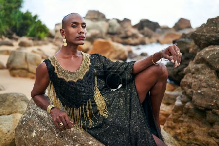 Photo for Epatage lgbtq black man poses on a scenic ocean beach looks at camera demonstrates jewellery. Androgynous ethnic fashion model in a posh dress, jewelry looks at camera, sits on stone. Pride month. - Royalty Free Image