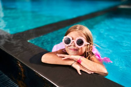 Kid relaxes on edge, cool water on summer vacation. Smiling girl in pink swimwear, goggles enjoys pool time on sunny day. Happy childhood, leisure swim, outdoor fun, warm weather activities.