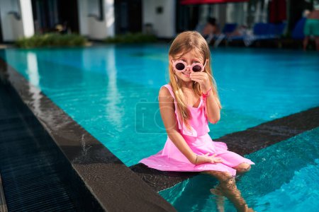 Child enjoys summer day, playful fashion, poolside fun. Little girl in pink dress, heart-shaped sunglasses sits at pool edge, feet dipped in water. Bright sunlight reflects on turquoise surface.