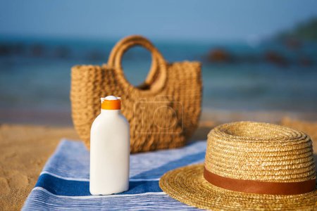 Summer skin care essentials by the sea. Bottle of high SPF sunscreen, straw hat, on towel at sunny beach. UV protection items on sand. Holiday accessories, sun cream ready for sunbathing by ocean.