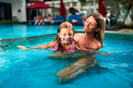 Summer day, family fun, mom teaches daughter swim strokes, outdoor luxury resort background, vacation activity. Smiling young girl in swimsuit learns swimming in pool, guided by her attentive mother.