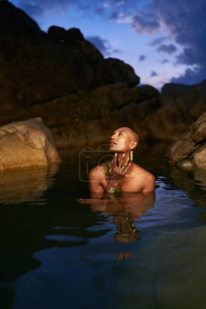 Persona negra no binaria se encuentra en el pecho profundo en medio de agua quieta dentro de un arroyo pintoresco por la noche. Lgbtq modelo de moda elegante étnica posa inmerso en un estanque cristalino en un lugar exótico en