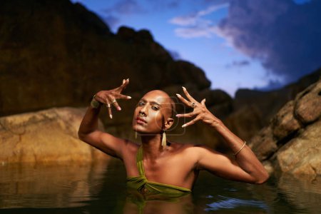Non-binary person of color poses in water, shows jewelry inside scenic creek at night. Lgbtq ethnic graceful fashion model wear brass rings with gems, nose-ring, earrings, bracelets immersed in pond.