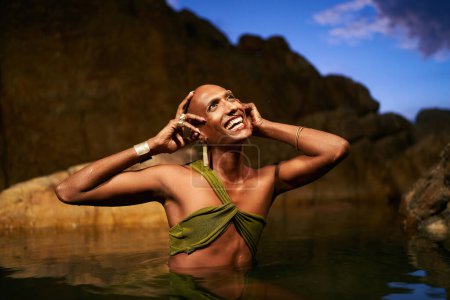 Lächelnde Geschlechtsflüssigkeit schwarze Person posiert in natürlichen stillen Wasser Pool. Queer Ethnic Fashion Model in offenem Kleid, Messingschmuck mit Edelsteinen, die in der Nacht anmutig inmitten eines felsigen Sees stehen.