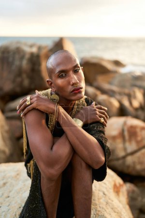 Photo for Non-binary bipoc fashion model in dress, brass jewelry sits on rocks by ocean. Trans sexual black person with rings, nose-ring, bracelets, earrings in posh clothes poses in tropical seaside location. - Royalty Free Image