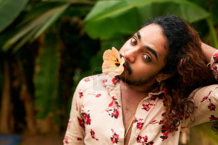 Flamboyant gay individual explores femininity, cultural identity through bold fashion, natural backdrop. South Asian man with expressive eyes, flower in mouth, models in tropical setting.