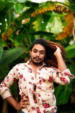 He poses confidently, hand in hair, bold fashion, expression of LGBTQ pride and joy in nature backdrop. Vibrant man with rich curly hair, floral shirt stands in tropical green plants.
