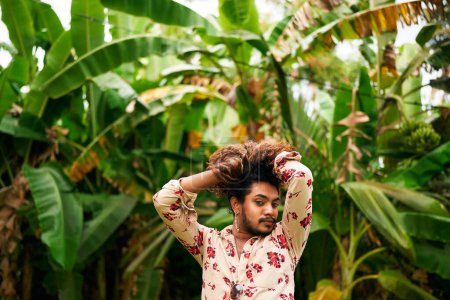 Confidence radiates as he touches voluminous hair, his gaze strong. Expressive gay man with floral shirt strikes playful pose amidst rich plants. Ideal for themes of LGBTQ pride, fashion.