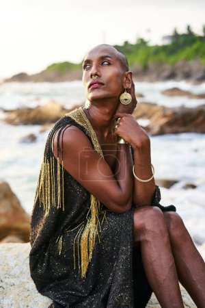 Nicht-binäre Bipoc-Modell in Kleid, Messing Schmuck sitzt auf Felsen am Meer. Transsexuelle schwarze Person mit Ringen, Nasenring, Armbändern, Ohrringen in nobler Kleidung posiert in tropischer Küstenlage.