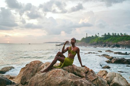 Gender flüssige schwarze Person posiert anmutig auf Felsen im Ozean bei Sonnenuntergang sitzend. Androgynes schlankes Modell ethnischer Mode in tropischem Maxi-Kleid auf einem felsigen Hügel über dem Sturm in der Abenddämmerung. Stolz auf LGBTQIA