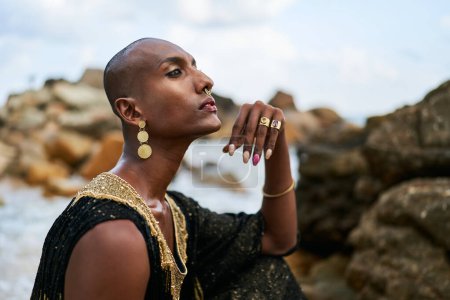 Photo for Epatage lgbtq black man poses on a scenic ocean beach looks at camera demonstrates jewellery . Androgynous ethnic fashion model in a posh dress looks at camera, sits on stone in - Royalty Free Image