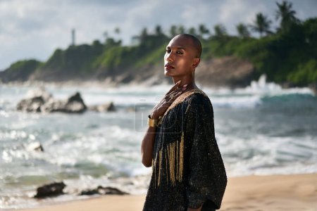 Photo for Outrageous lgbtq black model in luxury gown poses on scenic ocean beach with moody fashion look. Non-binary ethnic model in long posh dress and accessories looks at camera arrogantly, touches a neck. - Royalty Free Image