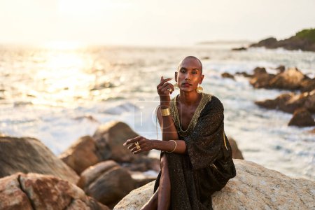 Non-binary black person in luxury dress, golden jewelry on beach rocks in ocean. Trans ethnic fashion model wearing jewellery in posh gown poses gracefully in tropical seaside location on a sunset.