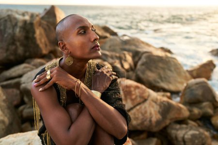 Queer black person in luxury dress, jewelry sits on rocks in an ocean. Lgbtq ethnic fashion model wearing jewellery dressed in posh gown poses gracefully in tropical seaside location. Pride LGBTQIA