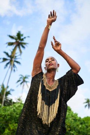 Epatage lgbtq mâle noir posant les mains en l'air sur la caméra sur la plage pittoresque de palmiers. Modèle de mode ethnique non binaire en robe longue chic porte des bijoux se tient gracieusement sur un emplacement tropical vert.