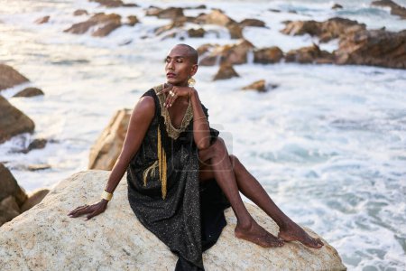 Non-binary bipoc fashion model in dress, brass jewelry sits on rocks by ocean. Trans sexual black person with rings, nose-ring, bracelets, earrings in posh clothes poses in tropical seaside location.