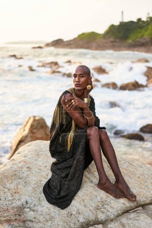 Queer black person in luxury dress, jewelry sits on rocks in an ocean. Lgbtq ethnic fashion model wearing jewellery dressed in posh gown poses gracefully in tropical seaside location. Pride LGBTQIA