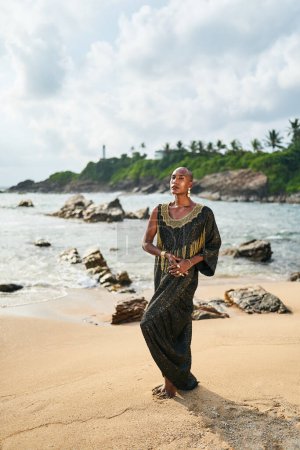 Empörender schwuler Schwarzer im Luxuskleid posiert am malerischen Strand des Ozeans. Nicht-binäre ethnische Mode Modell in schickem Outfit mit Make-up steht anmutig an malerischer Lage mit Meer, Leuchtturm.