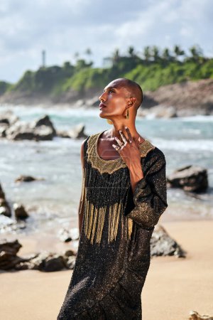 Trans sexual ethnic fashion model in long posh dress and accessories in elegant posture touches neck. Epatage gay black man in luxury gown poses, touches face on scenic an ocean beach. Pride month.