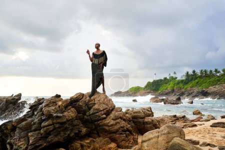 Une personne noire non binaire pose gracieusement debout sur des rochers dans l'océan. Modèle de mode trans ethnique dans une robe chic et des bijoux sur la plage rocheuse par la tempête. Lgbtq. Mois de fierté