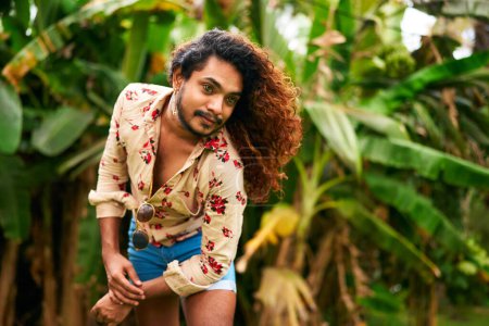 Expressive pose in lush plants, inclusivity and pride themes embodied. Confident gay man with curly hair in tropical setting poses with style, ethnic LGBTQ individual in floral shirt, denim shorts.