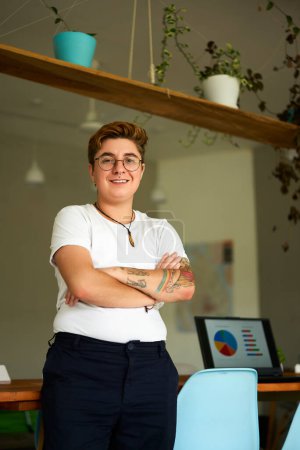 Inclusive workplace, gender identity expression. Transgender pro in modern office smiles confidently, crossed arms. Casual work attire, adorned with tattoos, of personal journey, empowerment.