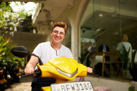Joyful moment, modern work-life balance with transport, represents diverse lifestyles. Transgender individual smiles atop yellow scooter outside coworking space, embracing urban eco commute.
