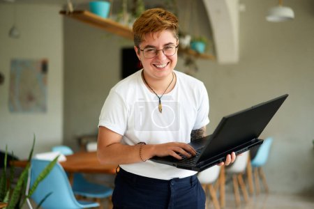 Inclusive workplace, young trans person enjoys career, experiences job fulfillment with tech in eco-friendly area. Smiling transgender professional holds laptop, works happily in modern office space.