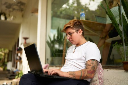 Relaxed office environment, gender diverse inclusive scene. Transgender pro works on laptop outdoors, focused on task. Comfortable casual attire, sunny business setting, remote work.