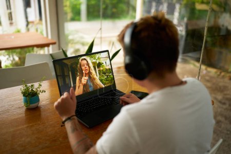 Young person with headphones communicates online, discusses project with colleague. Gen Z pro in casual office setup participates in video call from home. Modern work from anywhere concept.