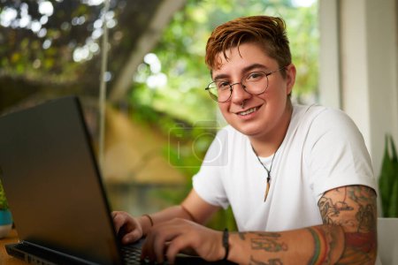 Photo for Pro in plant-filled room, reflecting modern, diverse work environments. Transgender male freelancer works at home on laptop, in casual white tee with tattoos, smiles at camera, workplace inclusion. - Royalty Free Image