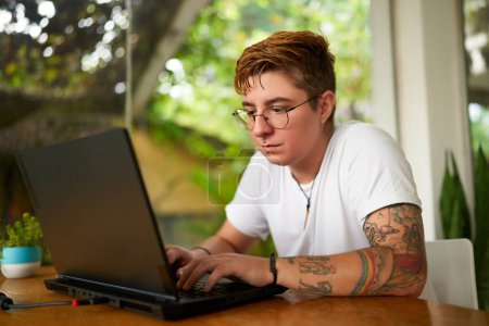 Focused individual with tattoos tech for remote job. Transgender freelancer works on laptop in tropical coworking space. Open, inclusive work plants. Creativity, productivity LGBTQ-friendly setting.