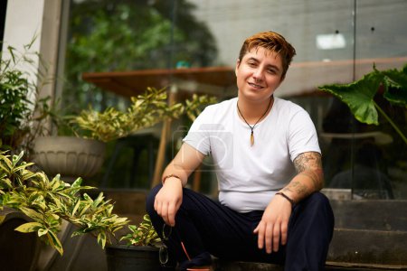 Inclusive workspace ambience with plants. Cheerful transgender person in casual wear works on laptop at co-working space. Digital nomad with tattoo enjoys remote job, smiles for camera in urban.