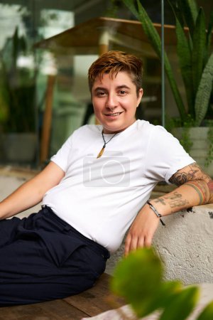 Smiling, tattooed trans man in white shirt enjoys break. Gen Z worker with tattoos sits casually in office environment. Inclusive workspace with modern decor. Young pro relaxes indoors.