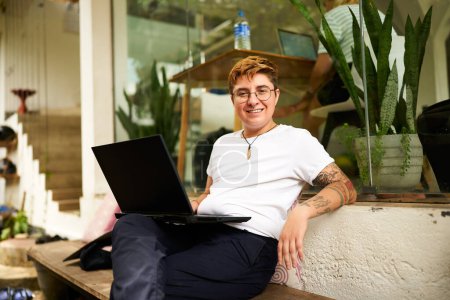 Inked, digital nomad in casual clothes, emphasizing diversity, inclusivity in modern remote work settings. Transgender Gen Z freelancer smiles while working on laptop at eco-friendly coworking space.