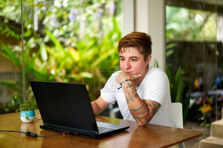 Confident and focused individual busy with work task, surrounded by green plants, embodying workplace inclusion and gender identity acceptance. Transgender pro works on laptop in home office setting.