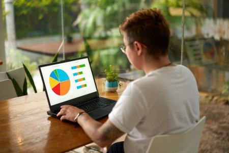 Short-haired worker in casual attire engages in remote work, data analysis, business strategy planning. Transgender professional analyzes pie chart, bar graph on laptop at eco-friendly office.