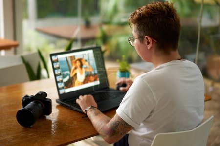 Tattooed, stylish person edits image, focused on screen, camera beside. Professional photo editor works on laptop in bright coworking space. Modern freelance job in creative industry, design workflow.