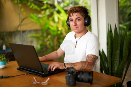 Focus, determination on face. Transgender editor with headphones edits video at laptop in casual office. Camera beside, rich plants background, inclusive workplace. Freelance, creativity, tech skills.