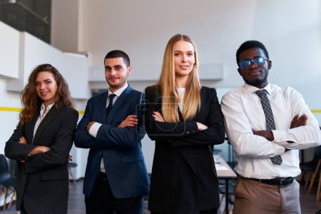 Caucasian female CEO with arms crossed flanked by diverse colleagues. Multiracial business team stands confidently in office. Professionals in suits portray unity, leadership in corporate setting.