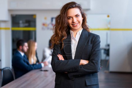 Colleagues in background hold a meeting. Confident businesswoman stands in office, arms crossed, smiling at camera. Professional, leadership, teamwork. Female executive, modern work environment.