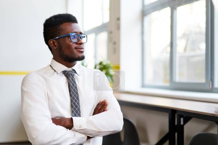Visionary leader in formal attire reflects diversity in workplace, success, leadership, growth. Confident African corporate pro stands in modern office, arms crossed, looking away optimistically.