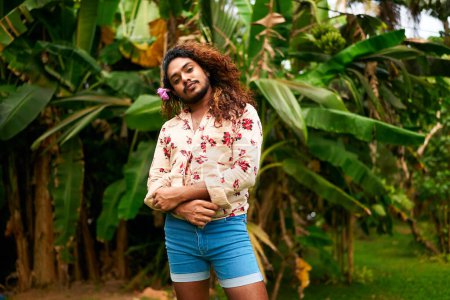 Wearing shorts and a floral shirt, he embodies pride and diversity, expressing his true self amidst nature. South Asian gay man with a flower in hair stands confidently in a rich garden.