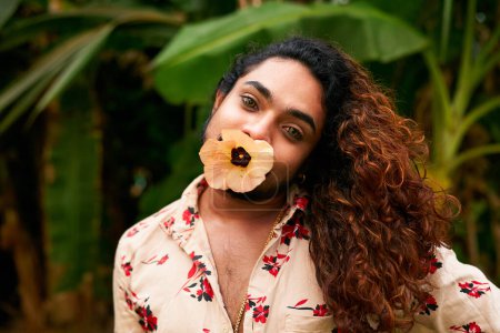 Wearing floral shirt amidst green foliage, represents gender fluidity, joyful confidence with tropical backdrop. South Asian man with flower in mouth poses, showing expressive makeup, bold femininity.