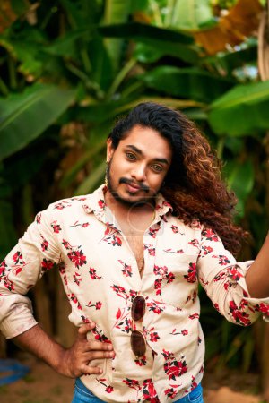 Hand on hip, looks directly, proud LGBTQ person with fierce expression. Confident gay man with wavy hair poses in tropical garden, vibrant floral shirt open, stylish sunglasses hang from collar.