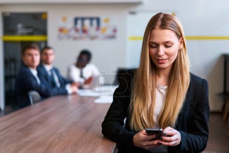 Professional multitasks on mobile, interacts with business apps. Blonde businesswoman in smart casual attire uses smartphone in office with colleagues in background. Modern communication at work.