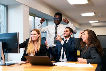 Multiethnic office team rejoices around computer, happy African exec raises fists in victory, coworkers cheer, man points at screen showing growth chart workplace celebration of successful deal.