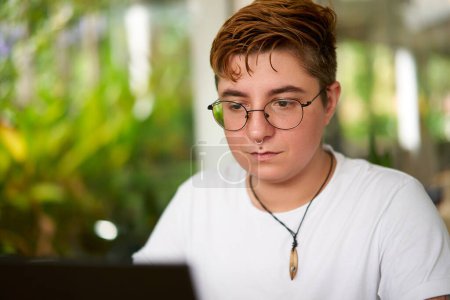 Inclusive environment, modern workplace diversity. Transgender pro focuses laptop work in office. Confident individual with glasses, casual attire. Background plants indicate eco-friendly space.