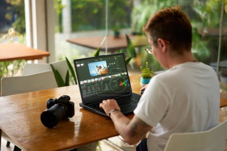 Color grading process by creative with camera gear beside. Pro videographer edits wildlife footage on laptop in a bright office. Concentrated freelancer tweaks travel video in a workspace.