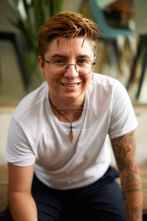 Confident diverse female to male with glasses, tattoos at work. Positive Gen Z transgender person smiles in coworking space. Young adult in casual wear, identity pride at modern office.